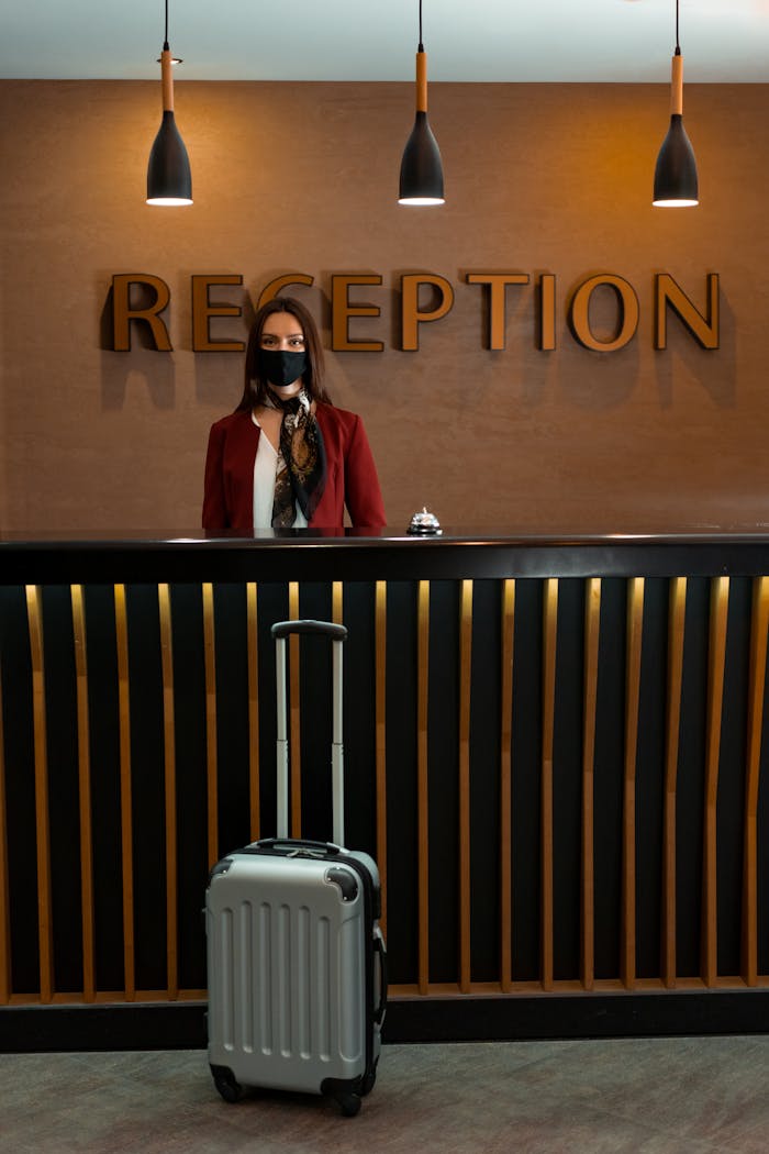 A hotel receptionist behind a desk wearing a face mask with luggage in front during the pandemic.